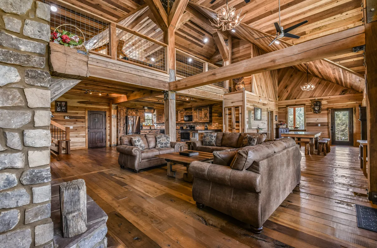 A welcoming, open interior featuring a stone fireplace, exposed wooden beams, plush sofas, and a full view of the adjoining kitchen area. The seamless flow between living and kitchen spaces encourages group interaction and highlights the central hub described in the section.