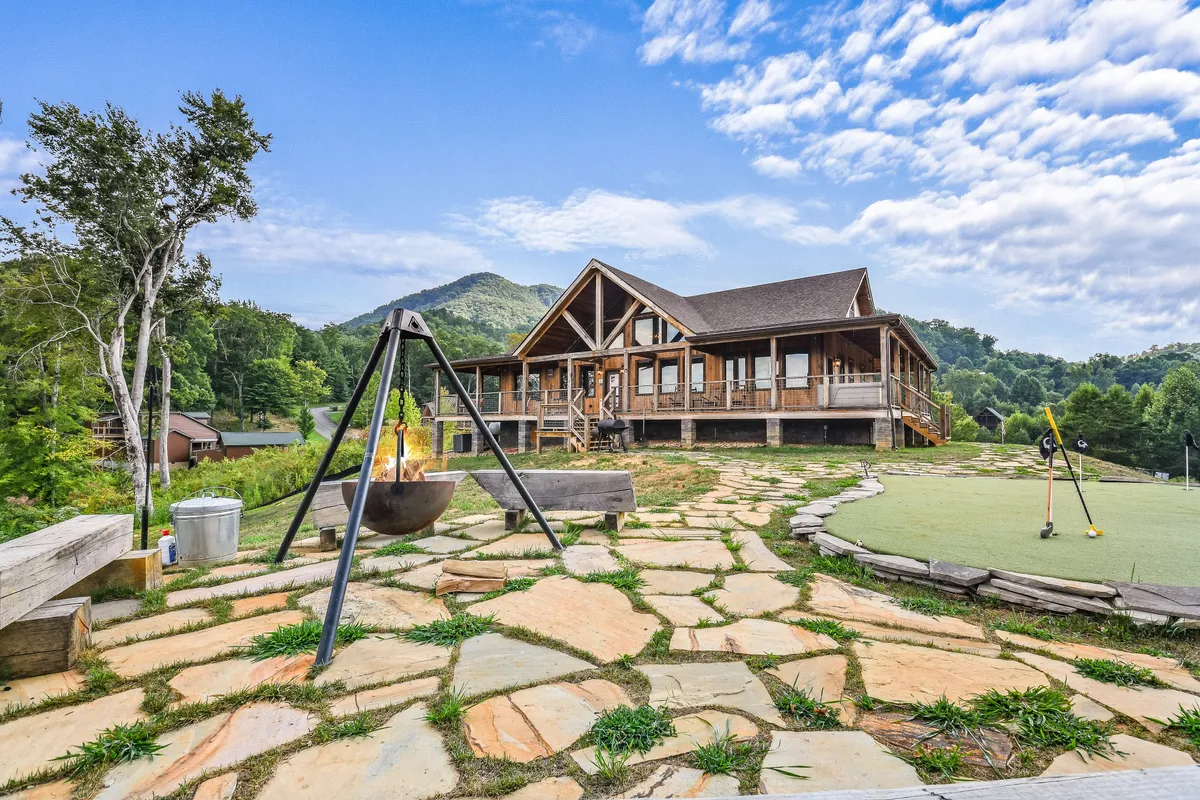 A spacious exterior scene showcasing a large wooden house, a fire pit surrounded by seating, a generous lawn for games, and a stunning mountain backdrop. The scene is designed for group activities and relaxed evening gatherings.