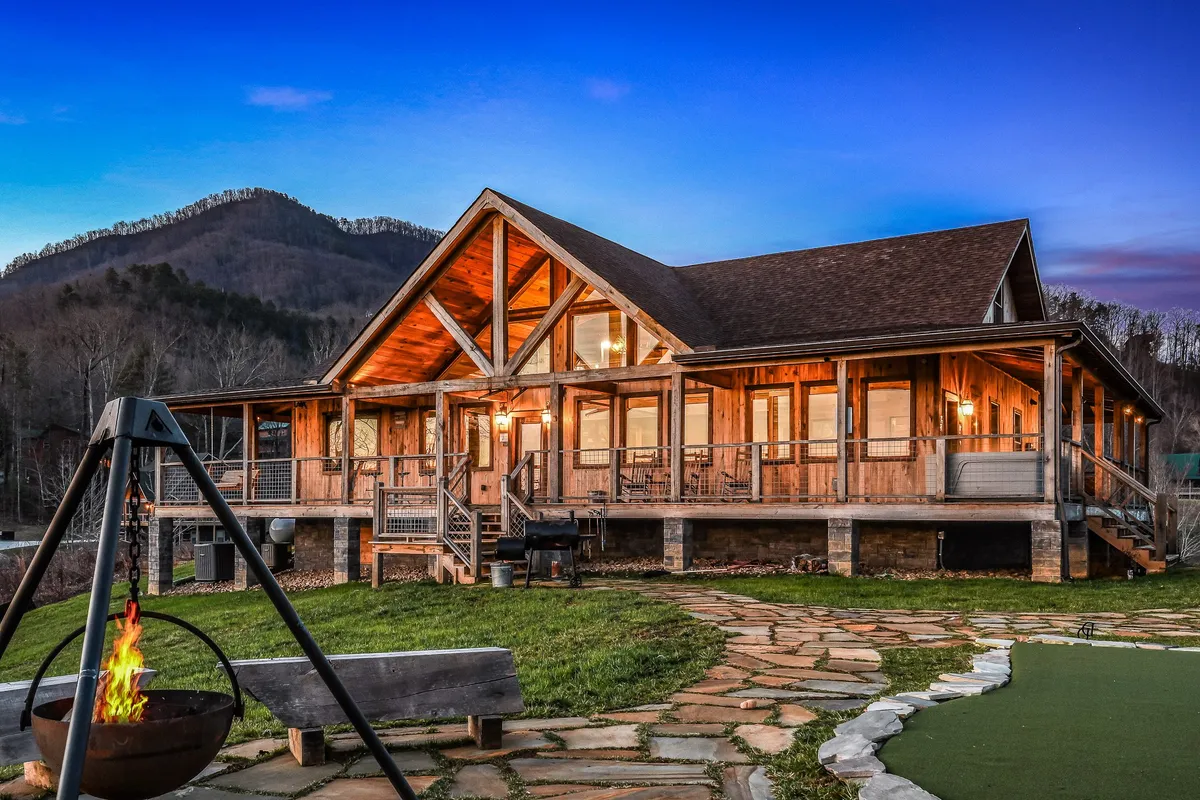 A spacious outdoor scene featuring a large wooden cabin, a welcoming stone pathway, a fire pit surrounded by seating, and a scenic mountain backdrop. The porch invites guests to relax, and the lawn provides space for games and gatherings. The setting is bathed in warm, cozy light, hinting at evening social activities.