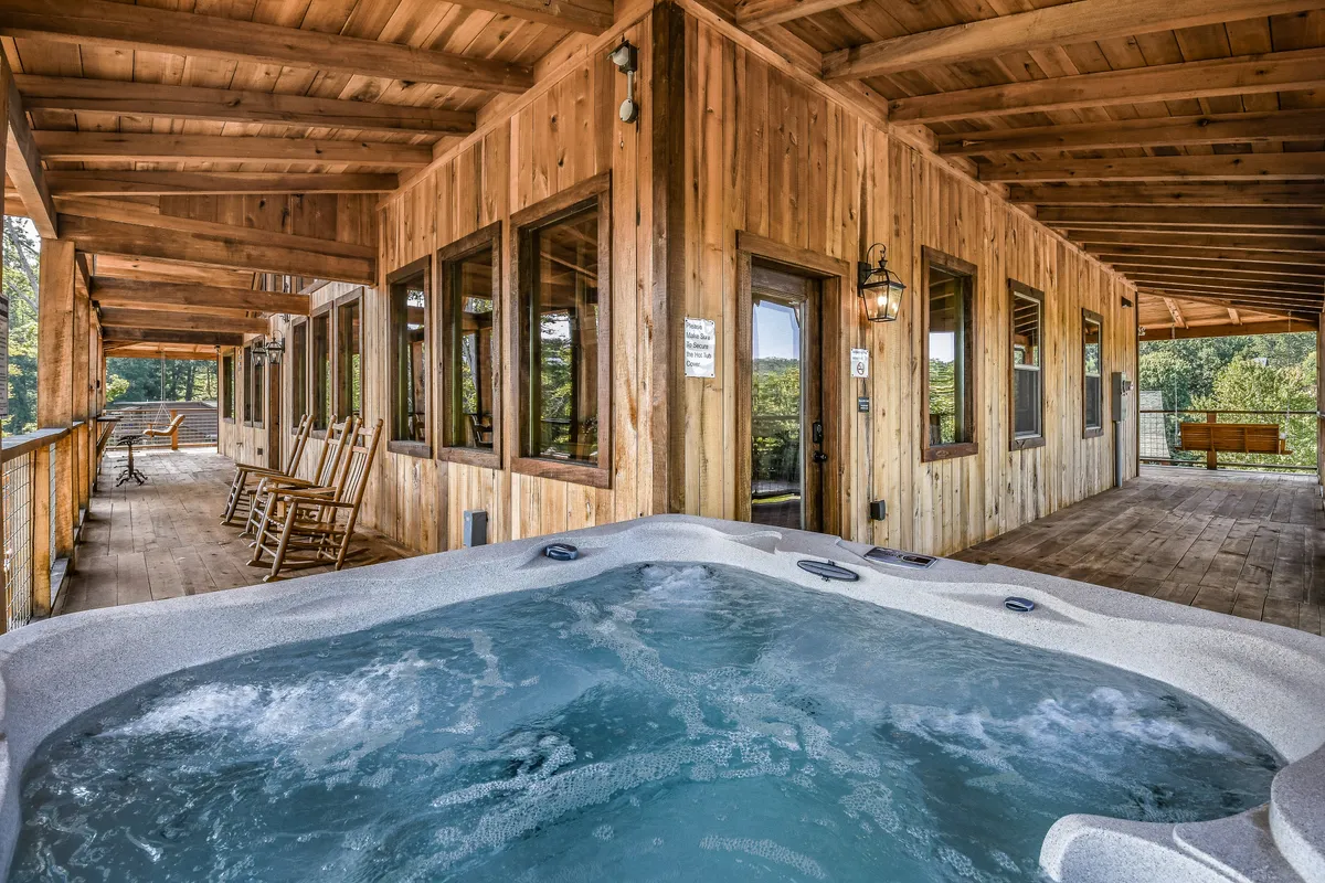 An exterior shot focuses on a wooden deck with a hot tub, surrounded by nature and large windows revealing the interior's inviting glow. Rocking chairs line the deck, offering a spot to relax and take in panoramic views, with the hot tub being the main feature for outdoor comfort and relaxation.