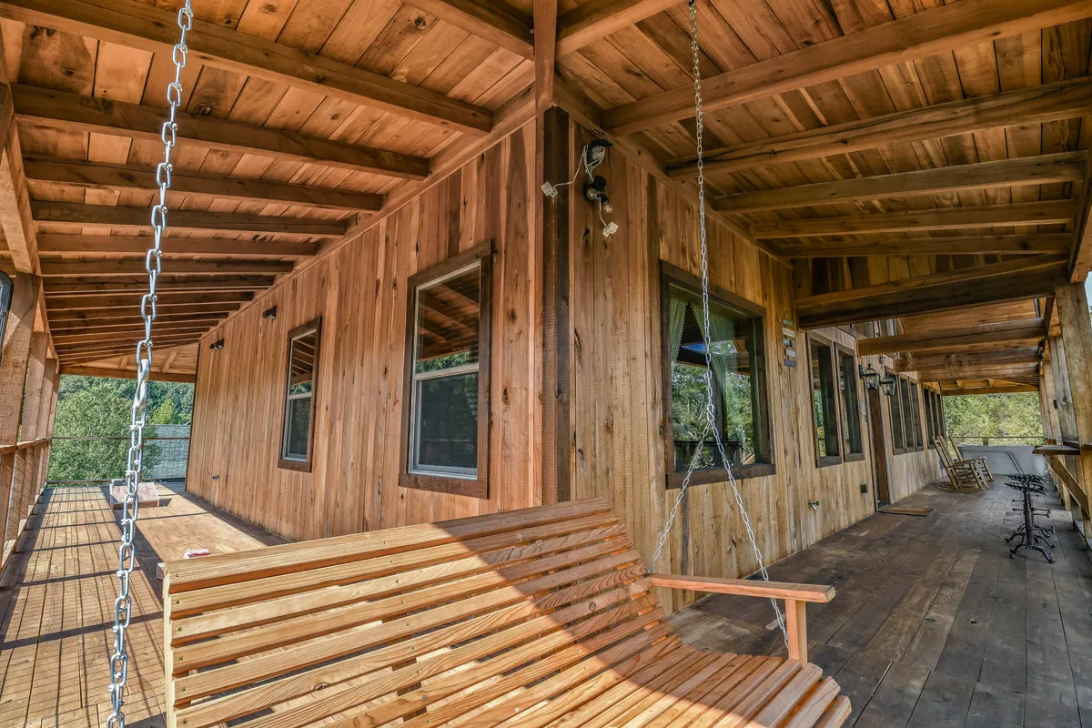 A peaceful porch scene features a wooden swing beneath exposed beams, with large windows offering glimpses inside and out. The view extends to the surrounding landscape, inviting guests to start their day with coffee or unwind as the sun sets.