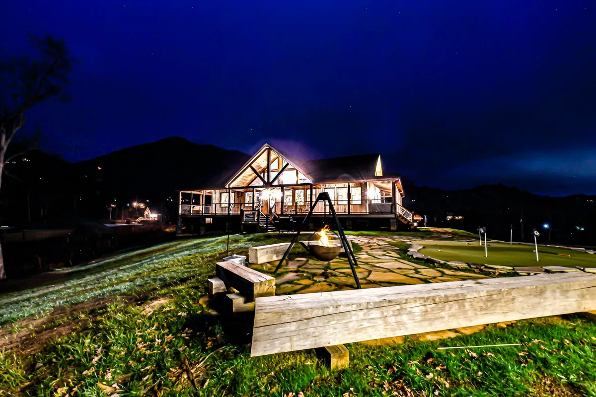An inviting evening shot of the large house, glowing warmly with interior lights. The foreground features a fire pit, a swing set, and a putting green, with the mountain skyline visible. The space feels lively and perfect for nighttime gatherings and games.