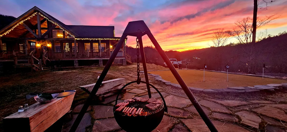 An inviting exterior shot of a wooden cabin with a grill loaded with food in the foreground, set against a dramatic sunset sky. The area is arranged for outdoor dining and group barbecues, with ample space for friends or family to play and relax.