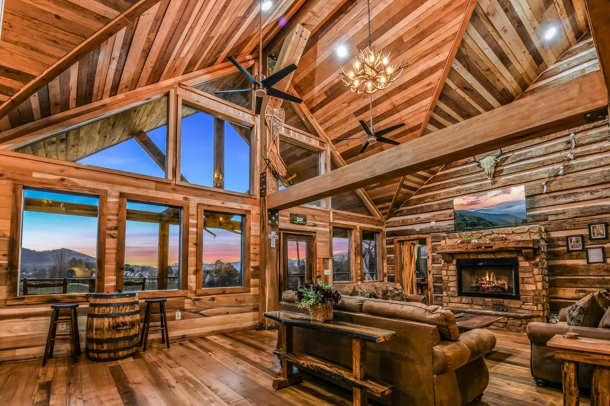This image showcases a spacious living area with large windows offering a sunset view, a prominent stone fireplace, rustic wooden beams, and comfortable, inviting furniture arranged for group gatherings. The space exudes warmth and offers an ideal setting for family gatherings, cozy evenings by the fire, and celebratory dinners.