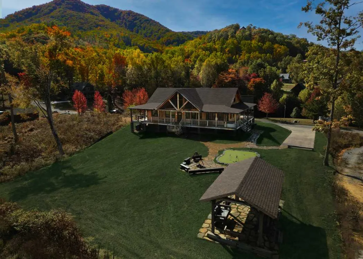 A wide exterior view of the luxury cabin shows the spacious lawn, surrounding mountains, and a prominent gazebo. The large wrap-around porch is visible, promising areas to relax and enjoy the outdoors. The setting is serene and welcoming, ideal for gatherings and taking in the scenic location.