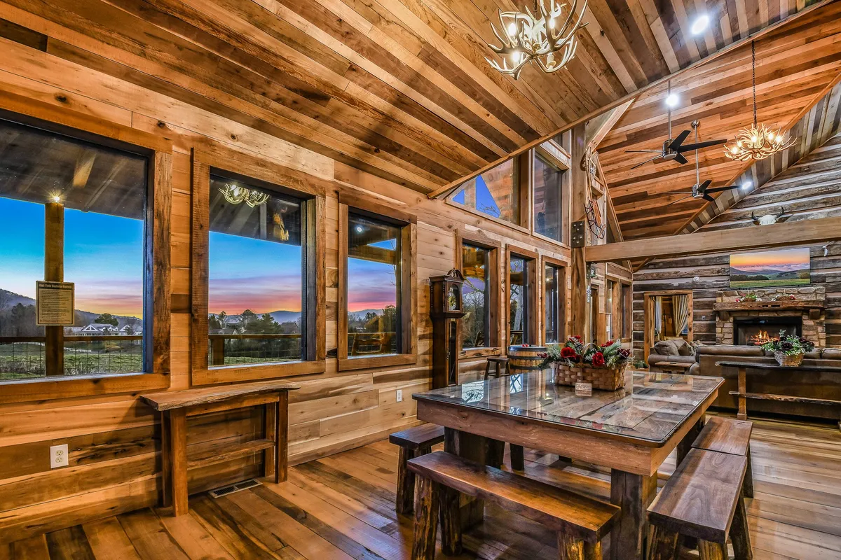 A large dining table sits before a glowing fireplace and expansive windows with a stunning sunset vista. Wooden beams add rustic charm, while the setup invites guests to gather for memorable meals or storytelling sessions.