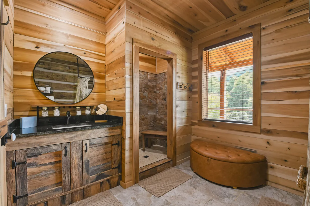 A stylish, tranquil bathroom with wooden cabinetry, a round mirror, a spacious walk-in shower, and an ottoman bench. Morning light illuminates the space, emphasizing the comfort and self-care amenities available for guests.