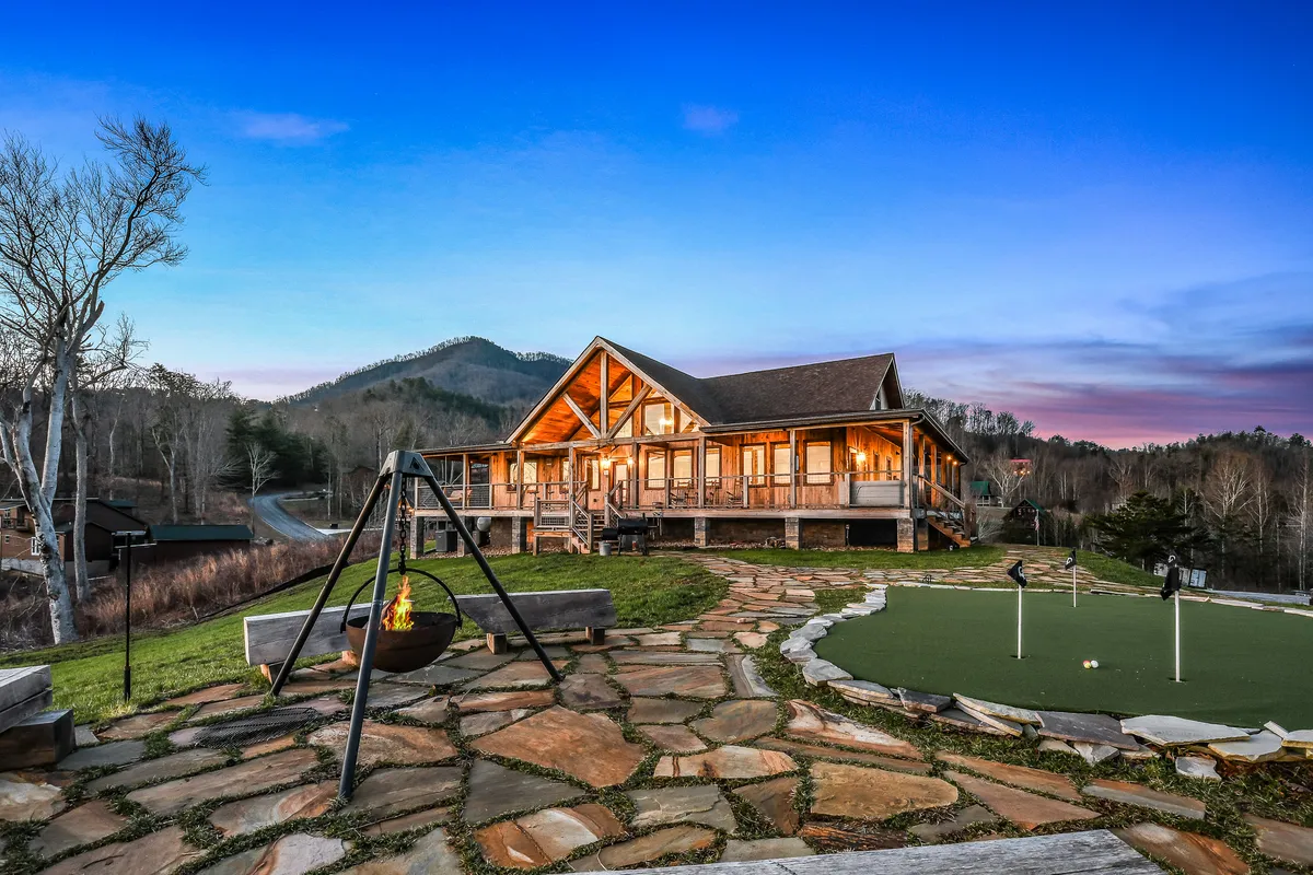 This exterior shot highlights a spacious cabin set against a backdrop of rolling mountains, featuring a cozy fire pit, putting green, and attractive stone pathways. The outdoor area is set up for gathering, relaxation, and play, with the mountains providing a dramatic context for the property.