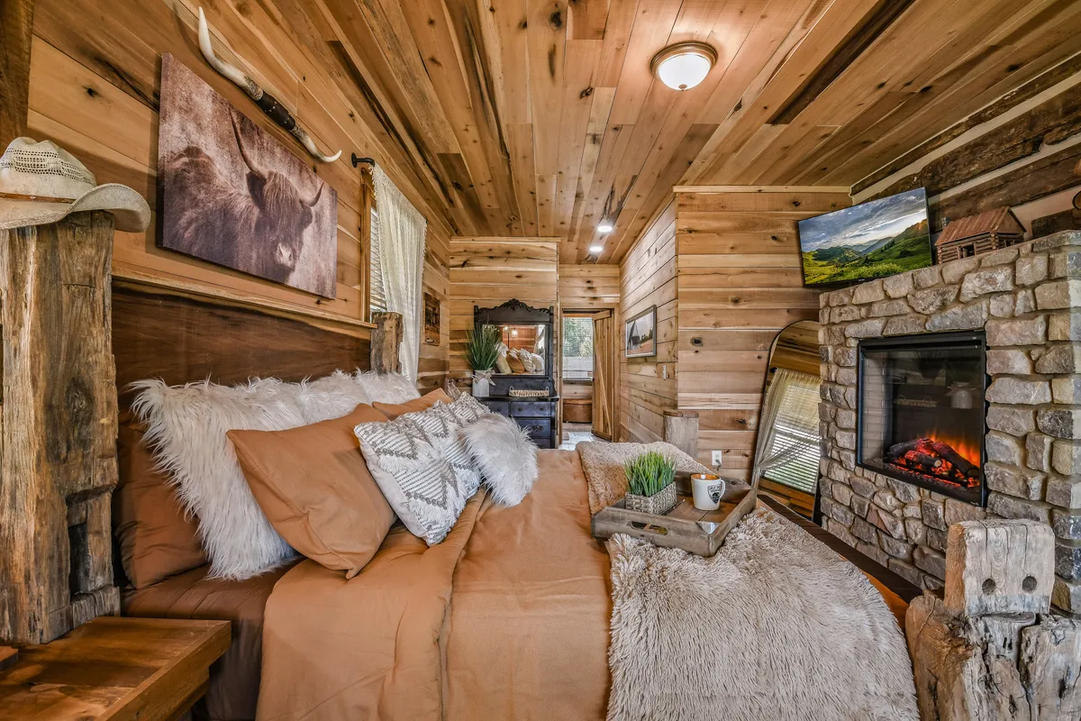 A cozy bedroom featuring a comfortable bed with fluffy pillows, a rustic stone fireplace, wooden accents, and tasteful artwork on the walls. Warm natural light filters through the windows, creating an inviting and restful atmosphere ideal for relaxation after a day of adventure.