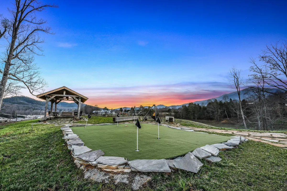 A vibrant green putting green is framed by a rustic shelter and set against a backdrop of mountains and a colorful sunset sky. The space is open and inviting, ideal for family golf games and outdoor gatherings.